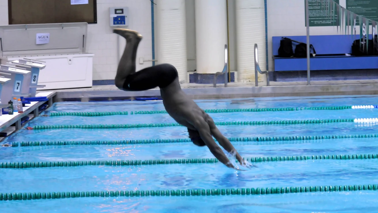 A swimmer dives into a pool, arms extended forward, at an indoor swimming facility.